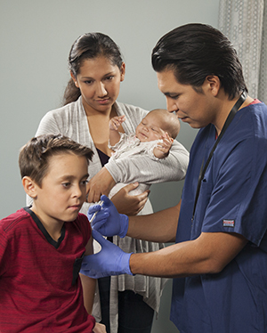 Health care provider giving boy an injection. Mother holding baby in background.